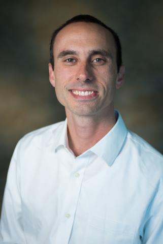 Andrew Culotta smiles at the camera, wearing a white button down shirt loosely buttoned at the collar.