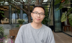 Young man in a grey t-shirt and glasses smiles at the camera, outdoors.