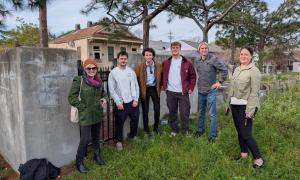 Six people stand in a grassy area in front of a low wall and a house.