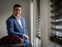 Michael Naguib in a blue suit leans on a leather chair next to a window.