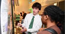 Man in a white shirt and green tie explains a poster to a woman in glasses.