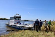 People disembark a boat on a grassy shore under a blue sky.