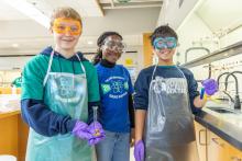 Three smiling students in a lab, wearing goggles and aprons, hold test tubes.