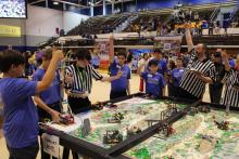 Students and referees celebrate near a detailed robot competition table.
