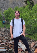 Smiling man with glasses and a backpack on a rocky, wooded trail.