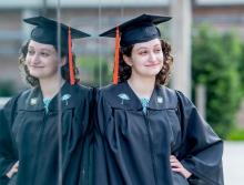 Young woman in graduation cap and gown smiles, her reflection visible.