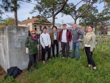 Six people stand in a grassy area in front of a low wall and a house.